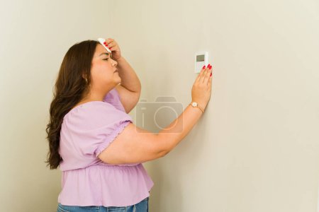 Téléchargez les photos : Femme inquiète se sentir chaud en été à la maison tout en allumant la climatisation à l'aide du thermostat et ayant des problèmes - en image libre de droit