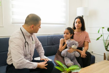 Photo for Scared little kid hugging her teddy bear while feeling sick and talking with a pediatrician during a doctor's visit - Royalty Free Image