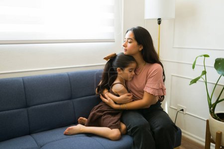 Photo for Beautiful worried mother hugging her sick little daughter looking sad while sitting together on the couch at home - Royalty Free Image