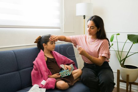 Téléchargez les photos : Triste maman hispanique à la maison prenant la température de sa petite fille malade avec une fièvre à l'aide d'un thermomètre - en image libre de droit