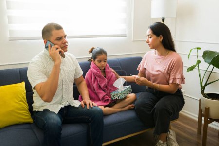 Photo for Hispanic dad and family calling the pediatrician on the phone for a doctor's visit because of his sick kid with a fever and cold - Royalty Free Image
