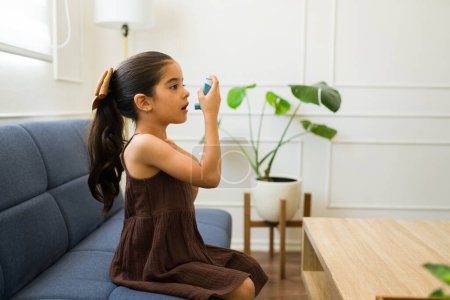 Téléchargez les photos : Adorable petite fille souffrant d'une crise d'asthme en utilisant un inhalateur pour aider à ses problèmes respiratoires seul à la maison - en image libre de droit