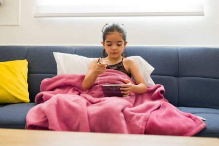 Photo for Beautiful sick latin 6 year old kid resting on the couch covered in a blanket while eating soup recovering from a cold or flu - Royalty Free Image