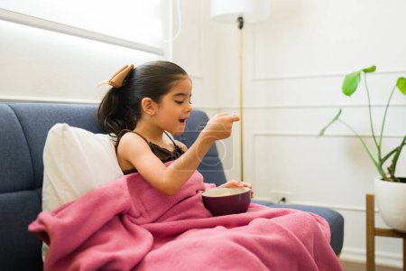 Photo for Adorable sick little girl eating hot soup while sick with a cold or flu resting on the couch living room - Royalty Free Image
