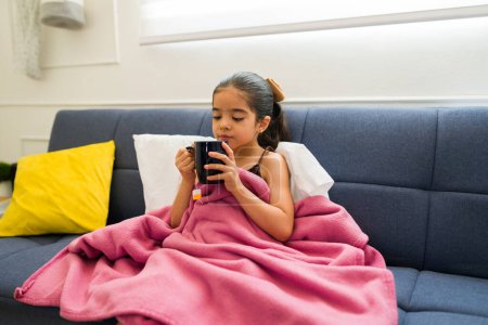 Téléchargez les photos : Petite fille mignonne enveloppée dans une couverture reposant sur le canapé se sentant malade en buvant du thé chaud récupérant d'un rhume ou de la grippe - en image libre de droit