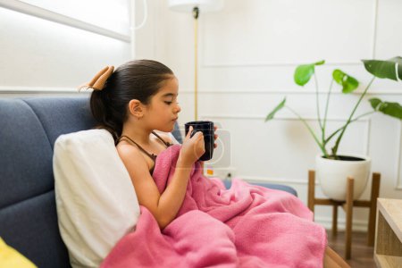 Photo for Beautiful hispanic young kid drinking hot tea with a blanket while recovering from a sick cold or flu at home - Royalty Free Image