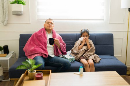 Photo for Father and daughter feeling ill and sick suffering from a bad cold or flu at home drinking hot tea and blowing their nose - Royalty Free Image