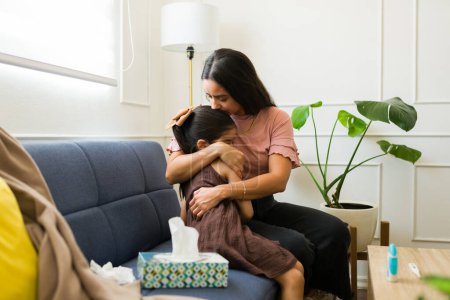 Téléchargez les photos : Belle jeune maman assise dans le salon embrassant sa fille malade de 6 ans tout en se sentant malade avec un rhume - en image libre de droit