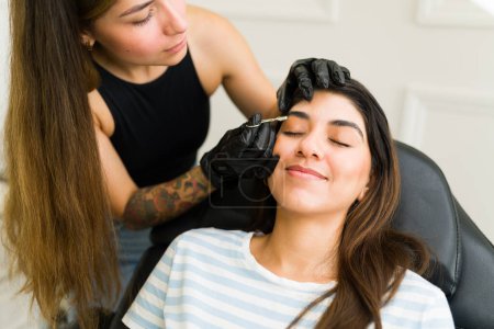 Téléchargez les photos : Esthéticienne utilisant une pince à épiler tout en faisant un nouveau design de sourcils sur une cliente heureuse au salon de beauté - en image libre de droit