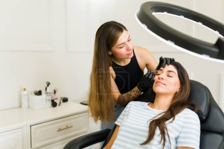 Photo for Beautician using tweezers making a new eyebrow design working with a happy hispanic woman client in the beauty room - Royalty Free Image