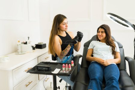 Foto de Hermoso cosmetólogo preparándose con un cliente feliz mientras prepara el pigmento de color para un servicio de tatuaje de labios - Imagen libre de derechos