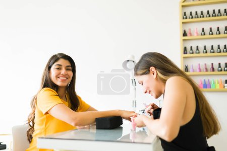 Téléchargez les photos : Femme mexicaine joyeuse souriant faire un contact visuel tandis que dans le salon de ongles peignant ses ongles avec de la couleur de gel après une manucure - en image libre de droit