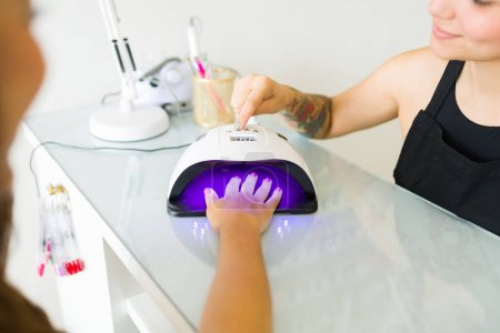 Téléchargez les photos : Femme gaie client dans le salon des ongles en utilisant l'éclairage UV après avoir peint ses ongles avec de la couleur de gel - en image libre de droit