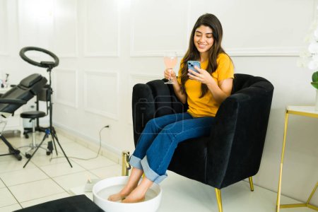 Foto de Relajada mujer joven feliz mensajes de texto en el teléfono inteligente mientras tiene un spa de pedicura relajante en el salón de uñas - Imagen libre de derechos