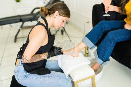 Foto de Hermosa trabajadora pintando las uñas de los pies de un cliente latino relajándose después de un relajante spa de pedicura - Imagen libre de derechos