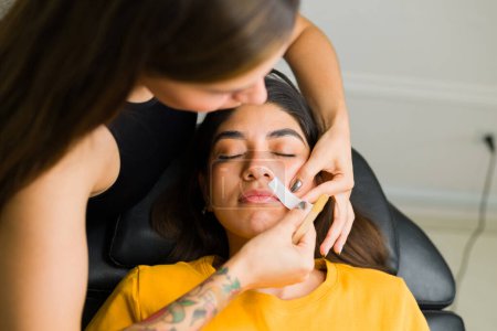 Téléchargez les photos : Esthéticienne latine utilisant des bandes de cire pour l'épilation sur la lèvre supérieure d'un client détendu dans la salle de beauté - en image libre de droit