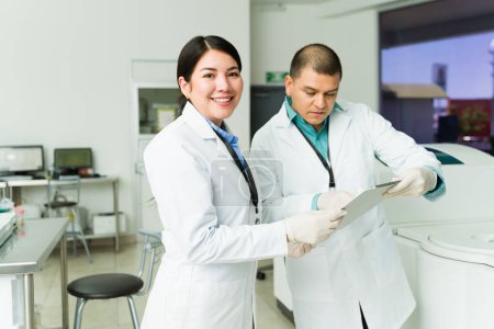 Téléchargez les photos : Belle femme heureuse chimiste souriant vérifier les résultats médicaux d'un test sanguin tout en travaillant dans le laboratoire - en image libre de droit