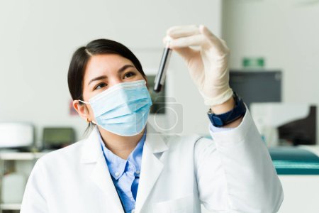 Téléchargez les photos : Heureux chimiste ou technicien caucasien avec masque facial et blouse de laboratoire regardant un échantillon de sang dans un tube à essai prêt pour un test médical - en image libre de droit