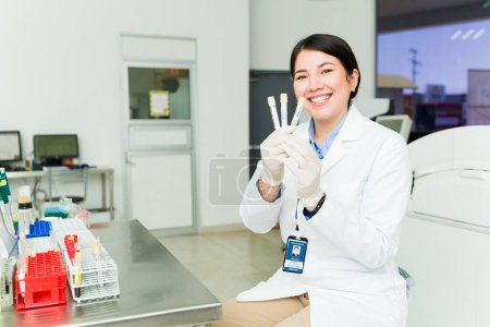 Téléchargez les photos : Joyeux beau chimiste souriant tout en tenant des éprouvettes et prêt à prélever des échantillons de sang dans le laboratoire médical - en image libre de droit