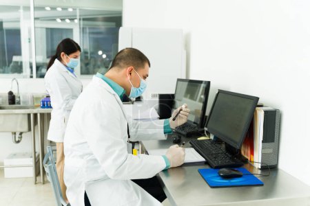 Foto de Hombre hispano y químico en la computadora sosteniendo una muestra de sangre en un tubo de ensayo mientras trabajaba en un laboratorio médico - Imagen libre de derechos