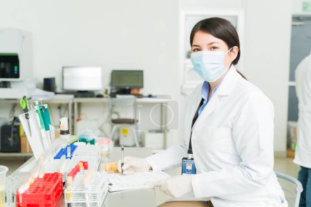 Photo for Beautiful happy woman with a face mask smiling working as a chemist or lab technician working test results of blood samples - Royalty Free Image