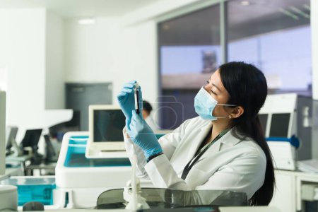 Téléchargez les photos : Chimiste caucasienne en laboratoire portant un manteau de laboratoire, des gants et un masque facial tenant une éprouvette avec un échantillon de sang - en image libre de droit
