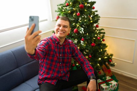 Photo for Attractive happy mexican man taking a selfie with her phone next to the christmas tree celebrating the holidays for social media - Royalty Free Image