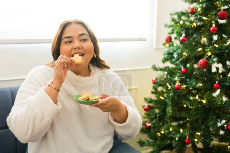 Photo for Cheerful attractive woman celebrating the holidays eating cookies at home next to her beautiful christmas tree - Royalty Free Image