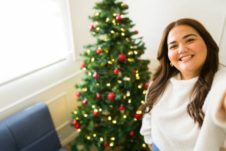 Photo for Personal perspective of a beautiful fat woman taking a selfie looking happy with her christmas tree celebrating the holidays at home - Royalty Free Image