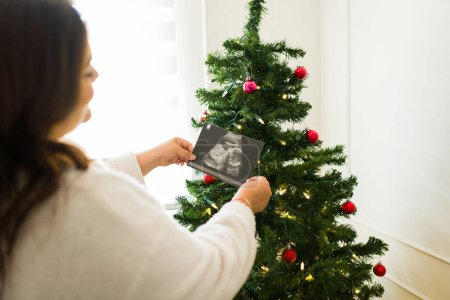 Photo for Latin plus size woman seen from behind putting a baby ultrasound on the christmas tree while happy about her pregnancy in the holidays - Royalty Free Image