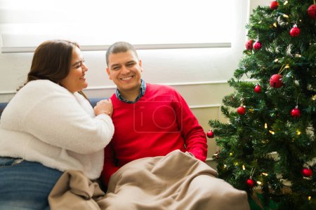 Photo for Happy beautiful couple looking relaxed at home laughing together using a blanket during a winter christmas celebrating the holidays - Royalty Free Image