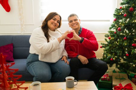 Photo for Attractive happy couple smiling making a heart gesture feeling in love while celebrating the christmas holidays together - Royalty Free Image