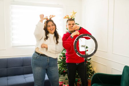 Foto de Atractiva pareja hispana con sombreros de reno bailando y filmando un video viral para las redes sociales usando una luz de anillo durante la Navidad - Imagen libre de derechos