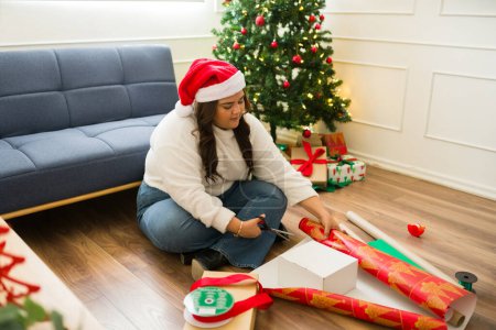 Foto de Mujer latina gorda con un sombrero de santa relajante y envolviendo regalos de Navidad para la familia para celebrar las fiestas - Imagen libre de derechos