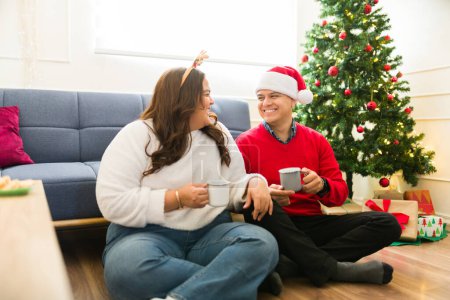 Téléchargez les photos : Grosse femme latine et homme heureux buvant ensemble du chocolat chaud et se relaxant dans le salon pendant la veille de Noël - en image libre de droit