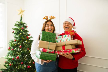 Photo for Excited couple with reindeer hats receiving or giving a lot of christmas presents smiling in front of the christmas tree at home - Royalty Free Image