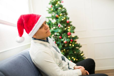 Foto de Triste latino de unos 30 años con un sombrero de santa se siente deprimido y solo celebrando la Navidad solo en casa - Imagen libre de derechos
