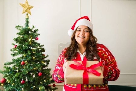 Téléchargez les photos : Belle femme heureuse de taille plus avec un chapeau de Père Noël donnant un cadeau et souriant en regardant la caméra devant l'arbre de Noël - en image libre de droit