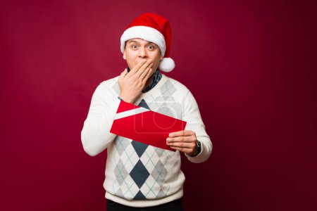 Téléchargez les photos : Surpris homme avec un chapeau de Père Noël couvrant sa bouche regardant surpris après avoir reçu une carte cadeau maquette pendant Noël - en image libre de droit