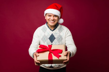 Téléchargez les photos : Un homme souriant dans la trentaine qui fait un contact visuel tout en offrant un beau cadeau de Noël portant un chapeau de Père Noël devant un fond rouge - en image libre de droit