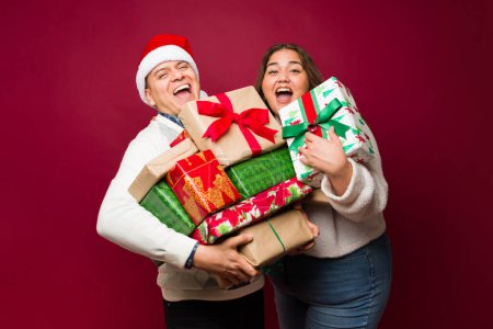 Foto de Feliz pareja hispana sonriendo y riendo mientras da o recibe muchos regalos de Navidad frente a un fondo rojo - Imagen libre de derechos