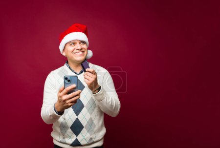 Téléchargez les photos : Heureux homme hispanique faire des achats en ligne sur son téléphone et payer avec une carte de crédit pour ses cadeaux pendant les vacances de Noël - en image libre de droit