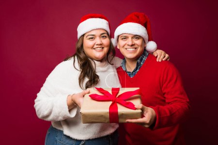 Photo for Attractive hispanic couple smiling while giving christmas gifts looking happy and hugging against a red studio background - Royalty Free Image
