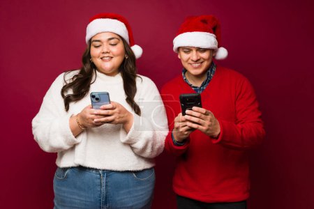 Foto de Feliz hermosa pareja sonriendo con sombreros de santa mientras que los mensajes de texto en su teléfono inteligente para celebrar la Navidad contra un fondo rojo - Imagen libre de derechos