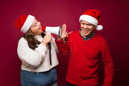 Photo for Plus size woman with a santa hat screaming about christmas gifts with a megaphone to an upset latin man during the holidays - Royalty Free Image