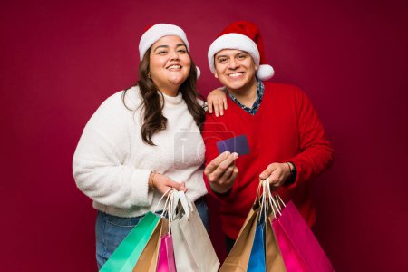 Téléchargez les photos : Beau couple hispanique avec chapeaux de Père Noël souriant avec beaucoup de sacs à provisions payant avec une carte de crédit pour leurs cadeaux de Noël - en image libre de droit
