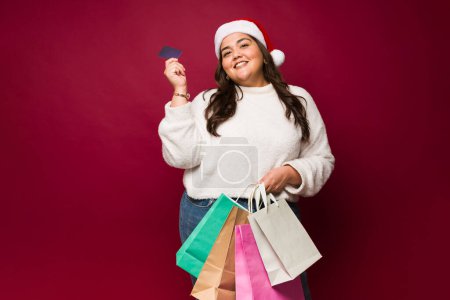 Téléchargez les photos : Mexicaine grosse femme souriant portant un chapeau de Père Noël sur un fond rouge faire du shopping pendant les vacances de Noël acheter des cadeaux - en image libre de droit