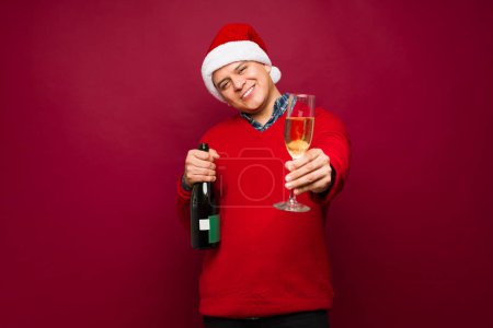 Foto de Hombre feliz en sus 30 con un sombrero de santa bebiendo champán y diciendo vítores celebrando la Navidad sonriendo - Imagen libre de derechos