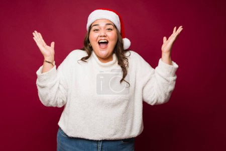 Photo for Overweight hispanic woman looking excited and surprised wearing a santa hat happy about the christmas holidays - Royalty Free Image
