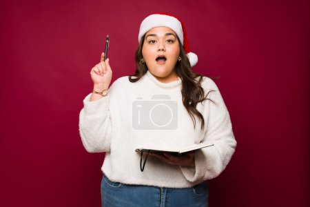 Foto de Mujer gorda hispana sorprendida mirando emocionada mientras escribía una carta de santa para sus regalos de Navidad o lista de compras en un fondo rojo - Imagen libre de derechos